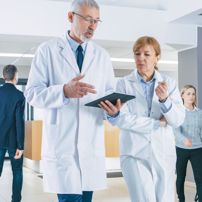 In The Hospital, Busy Doctors Talk, Using Tablet Computer While Walking Through The Building. New Modern Fully Functional Medical Facility.