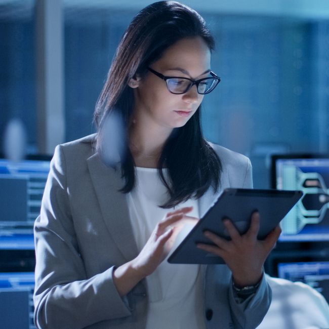 Young Female Government Employee Wearing Glasses Uses Tablet In System Control Center. In The Background Her Coworkers Are At Their Workspaces With Many Displays Showing Valuable Data.