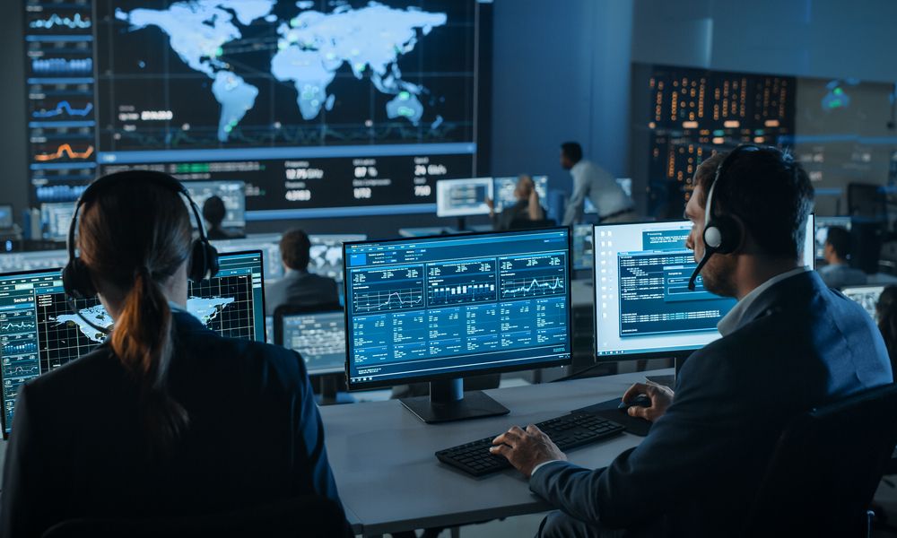 Shot Of Officers In A Surveillance Control Center With Police Global Map Tracking On A Big Digital Screen. Monitoring Room Employees Sit In Front Of Computer Displays And Analyze Big Data.