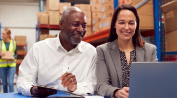 Male And Female Team Leaders Working On Laptop In Warehouse With Workers In Background