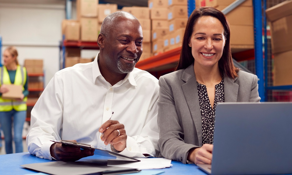 Male And Female Team Leaders Working On Laptop In Warehouse With Workers In Background